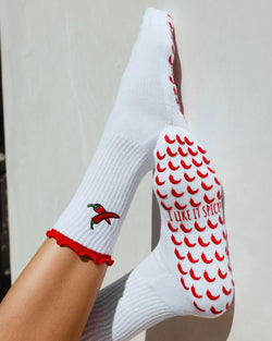 White sock with red chili pepper design and text held by a person against a neutral background. Pilates grip socks. 